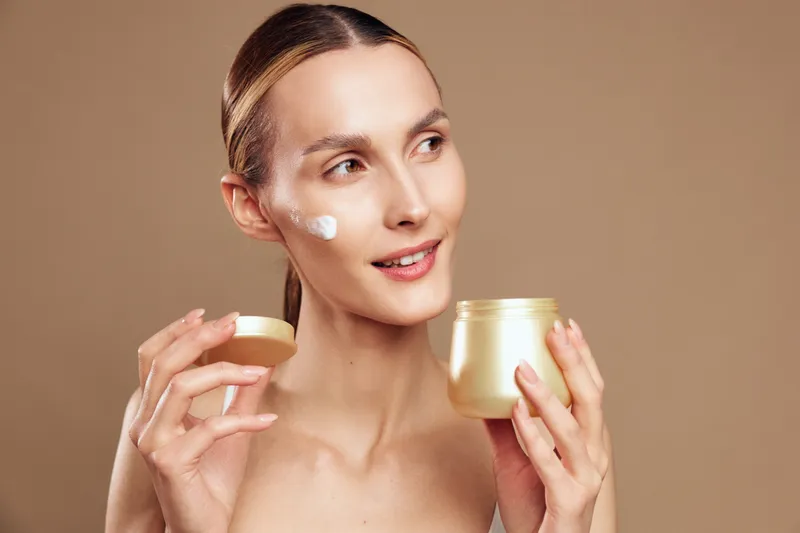 Young woman applies face cream with fingers holds open jar skincare product looks away smiling has clean skin natural beauty isolated on beige background.