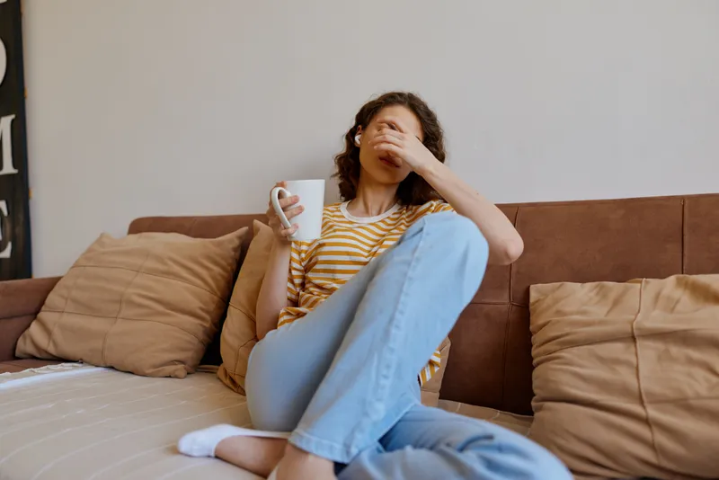 A stressed woman sits on a couch at home, head in hand, clutching a mug. The bright living room highlights fatigue, burnout, anxiety, and loneliness in a intimate moment.