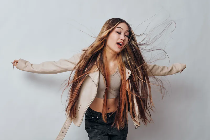 Young woman with long hair wearing a jacket and crop top dancing with eyes closed in front of gray background. Fashion, movement and joy with casual style and relaxed mood.