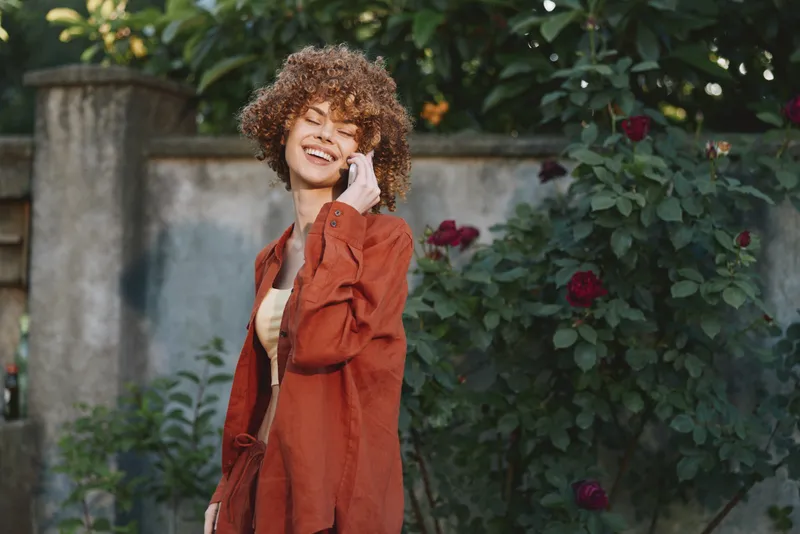 Cheerful woman outdoors, smiling while talking on phone, orange coat, casual lifestyle moment amid greenery and flowers, sunlit scene with relaxed posture