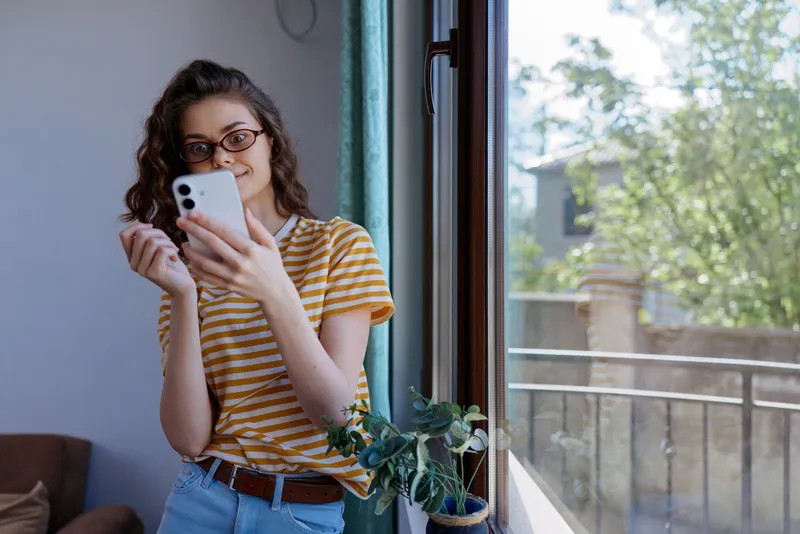 Woman in a yellow striped t shirt and jeans stands near an open window, smiling while scrolling a smartphone, indoor plant beside her, natural light creating a cozy home lifestyle scene.