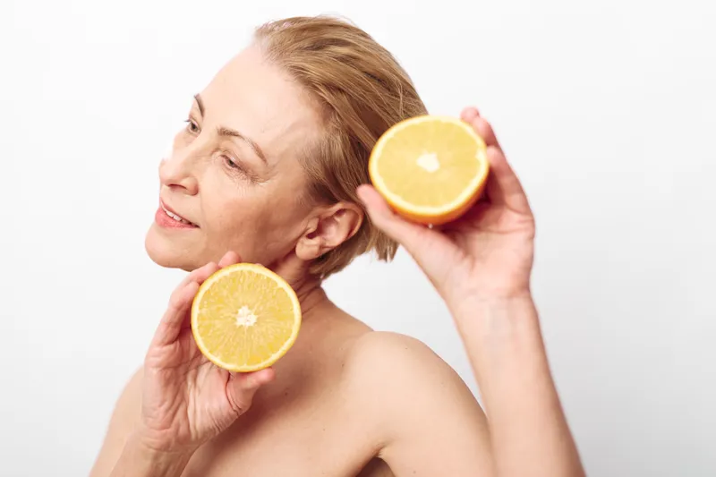 Mature woman with natural skin holds two orange halves near her face, smiling softly on white background. Concept of healthy lifestyle, skincare, nutrition, and natural beauty.
