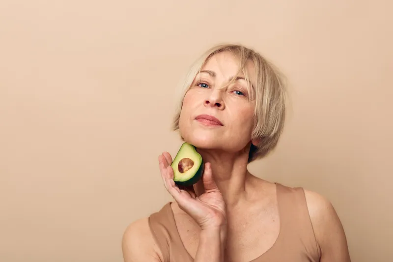 Mature woman with short blonde hair holds half an avocado near her face, posing confidently against beige background. Concept of healthy lifestyle, natural beauty, and wellness.
