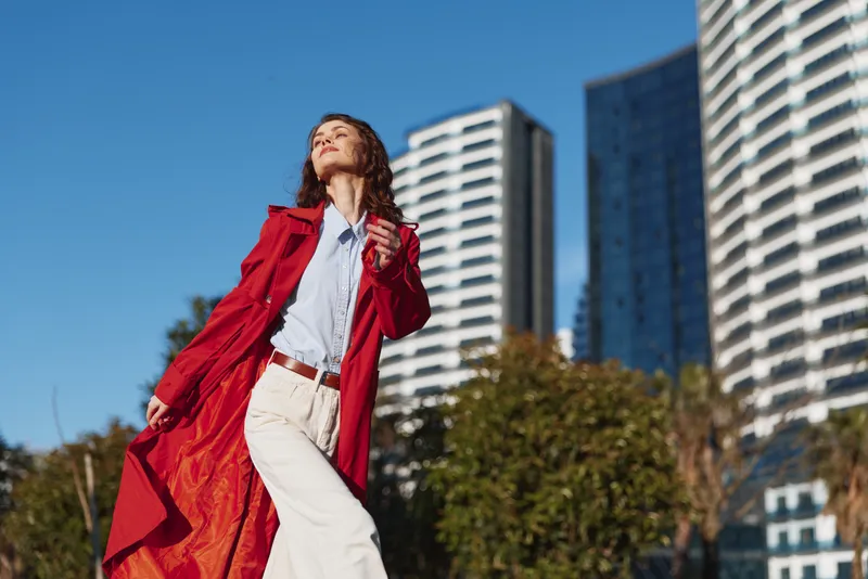 Young woman in a flowing red coat confidently walks outdoors in an urban setting with modern skyscrapers and clear blue sky in the background.