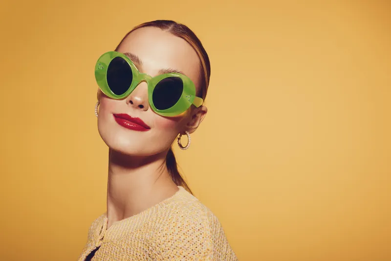 Young woman with slicked hair wearing large green sunglasses and red lipstick poses confidently against yellow background. Fashion concept with bold accessories and makeup style.