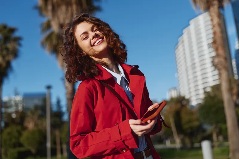 Young woman with curly hair wearing a red jacket smiles joyfully while holding a smartphone outdoors on a sunny day in an urban park setting.
