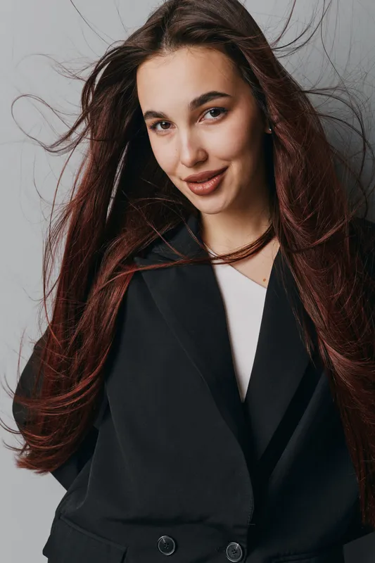 Young woman with long brown hair wearing black coat and white top posing confidently in studio. Female model with natural makeup and loose hair for fashion portrait and beauty concept.