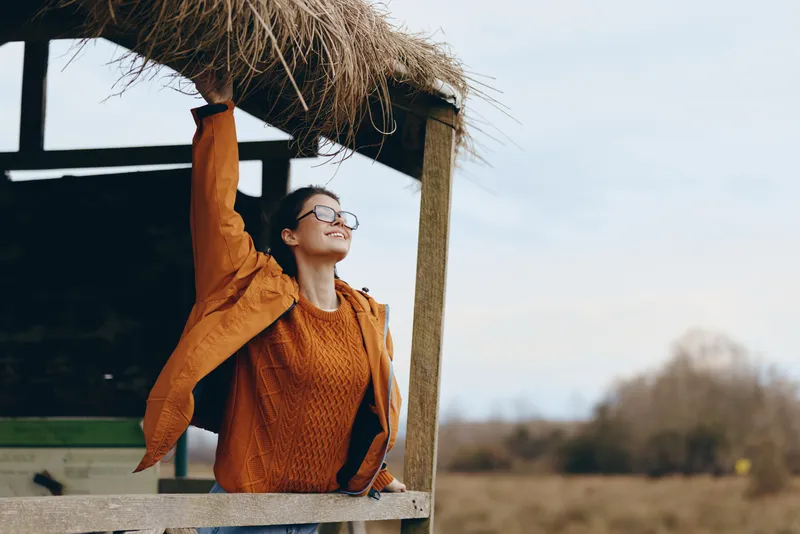 Woman lifestyle outdoor portrait, orange sweater and glasses on a terrace, she raises one arm and smiles at warm daylight sky, calm leisure mood with nature trees and soft wind in spring.