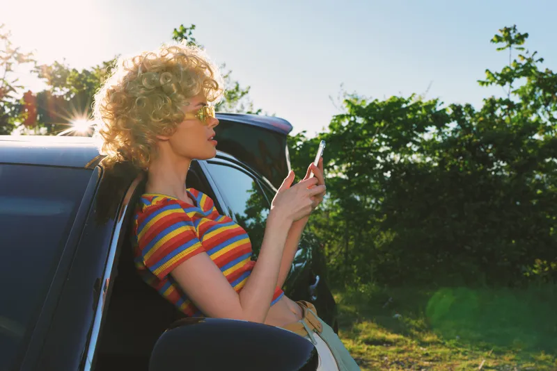 Joyful woman leaning from a car window, checking a smartphone, sunlit moment, retro color palette, casual striped top, outdoor calm, natural lighting