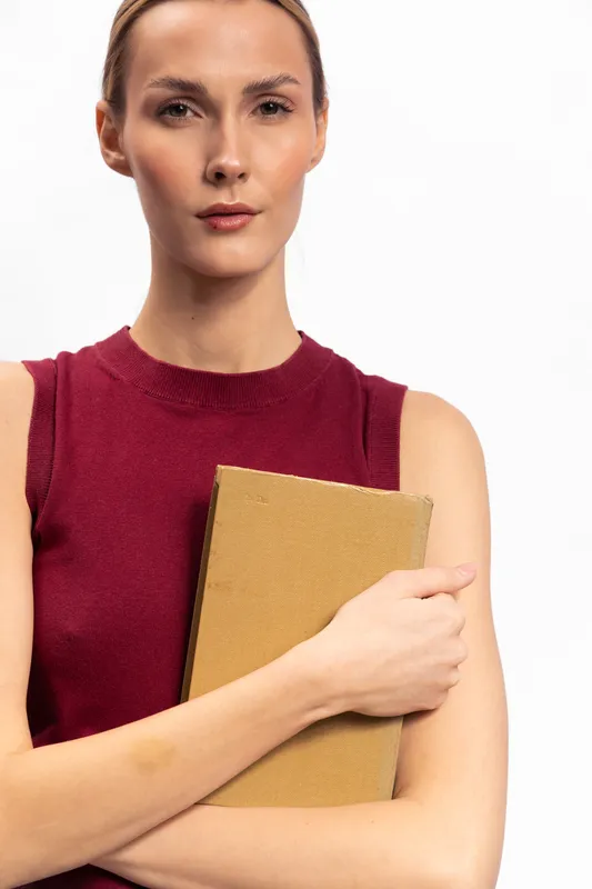 Confident young woman in sleeveless red top holds a closed book against white background. Studio portrait of serious female model with natural makeup and neat hairstyle, concept of knowledge.