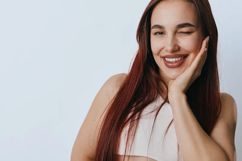 Young woman with long hair smiling and winking with hand on cheek against white background. Portrait of happy female with natural makeup and casual sleeveless top in studio lighting.