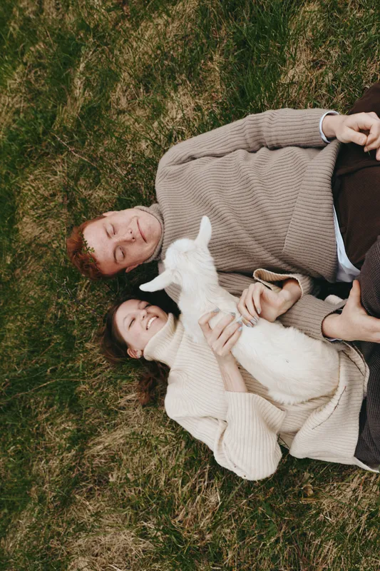 Two young women lying on grass with a white baby goat, enjoying a relaxed moment in casual sweaters during a peaceful outdoor lifestyle scene.