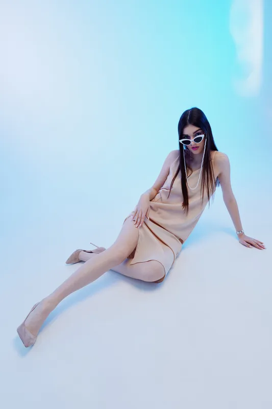 Young woman in a beige dress and sunglasses poses on the floor in a bright studio. Her long hair and stylish outfit emphasize beauty, fashion, and elegance concepts.