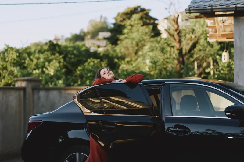 A smiling woman leans against a sleek car in a sunny driveway, embodying a casual, fashion forward lifestyle and confident pose in warm daylight