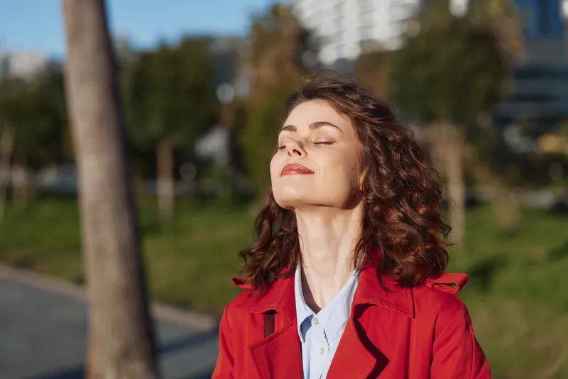 A young woman in a red coat enjoys the warm sun, eyes closed, feeling calm and free in a bright city park.
