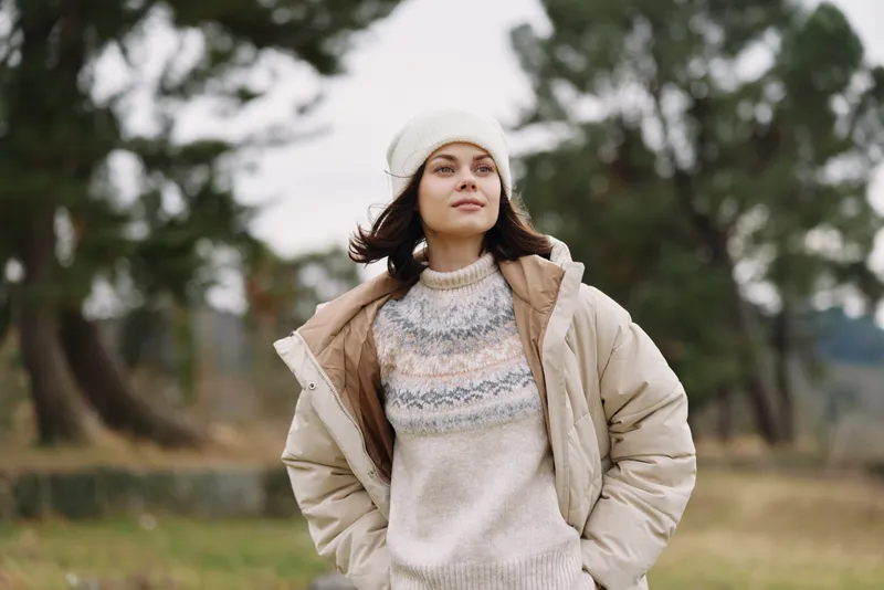 Woman in a beige coat and knit sweater stands outdoors in a park, wearing a knit beanie, conveying casual fashion and cozy warmth during a chilly day in nature