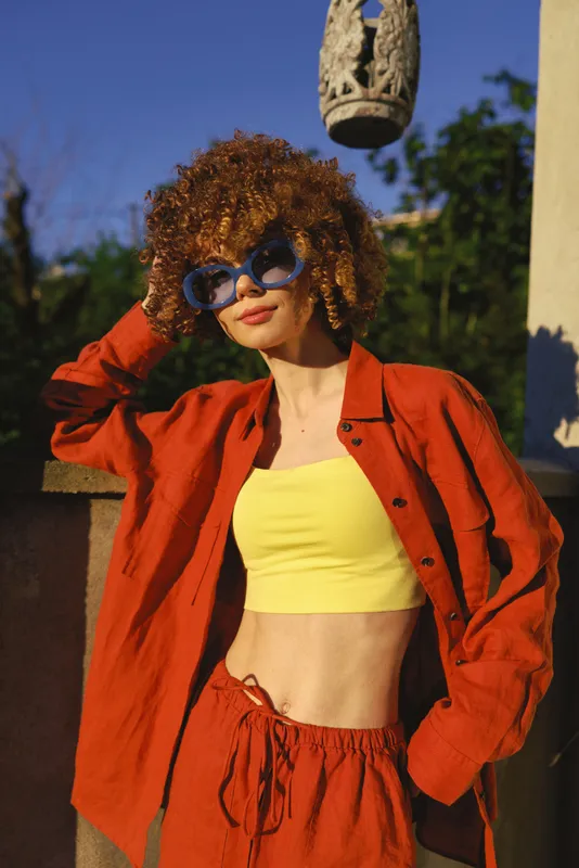 Young woman with curly hair poses outdoors in a bold red jacket and bright yellow crop top, wearing sunglasses and exuding confident street style