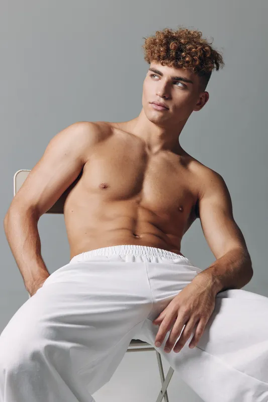 Young man with curly hair sitting shirtless on chair wearing white pants, looking away with relaxed posture in studio with neutral background and soft lighting.