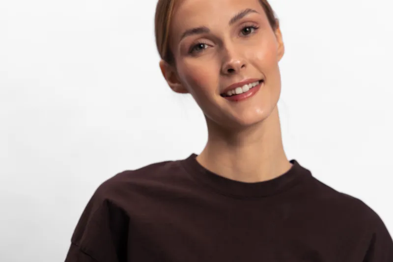 Smiling young woman with light skin and brown hair wearing dark shirt looks at camera on white background. Portrait of confident female model with natural makeup and positive expression.