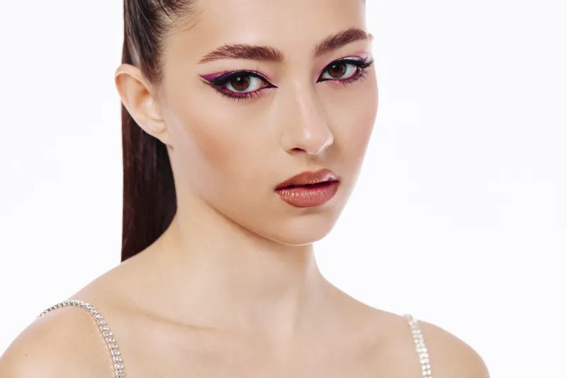 Young woman with artistic eye makeup and flawless skin poses against white backdrop. Portrait shows elegant fashion beauty look emphasizing creativity and style.