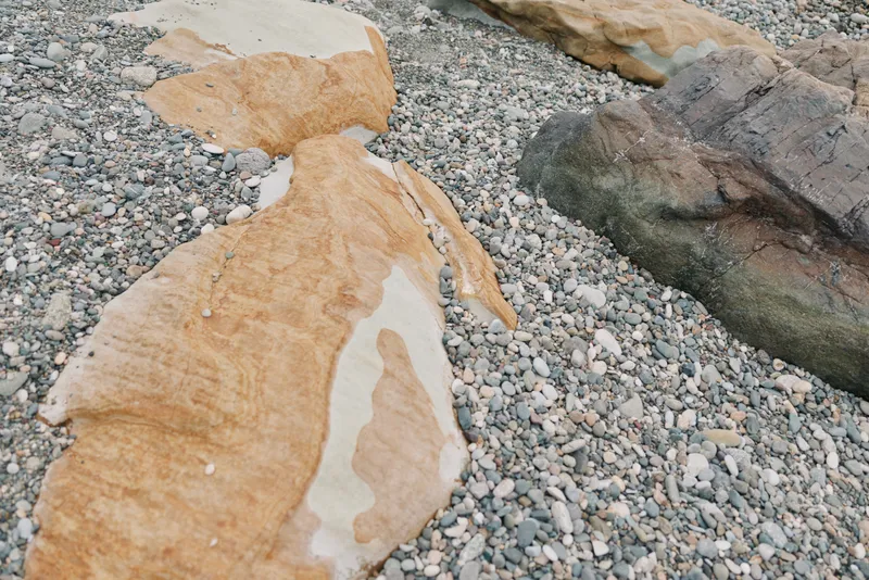 Large beige and brown rocks lie scattered on a pebble beach, showcasing natural textures and earthy colors under soft daylight. Concept of nature, geology, and coastal landscape.