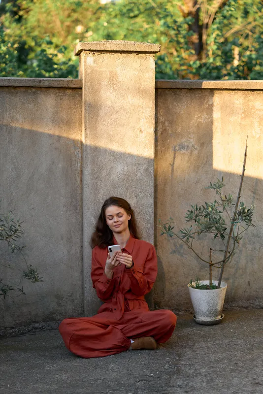 Woman sitting crosslegged in a red jumpsuit using a smartphone in an outdoor space, against a textured wall with a potted plant, conveying lifestyle, wellness, and daily tech use