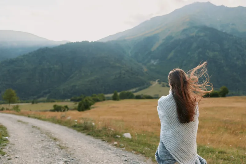 Woman standing outdoors in a open countryside with a winding path and distant mountains, wearing a cozy sweater, hair blowing, capturing a serene lifestyle moment