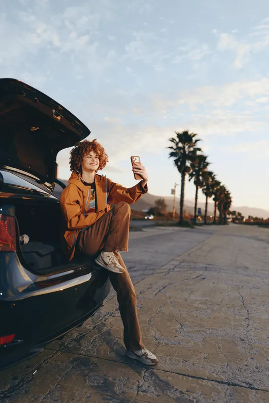 Woman lifestyle outdoors by car at sunset, smiling and holding phone near the open trunk, warm golden light, relaxed posture, curly hair, palm trees background, casual street