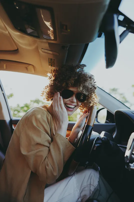 Smile of a woman with curly hair in a car, driver at the wheel, lifestyle moment with sunglasses, interior light and sunlight highlighting everyday joy.