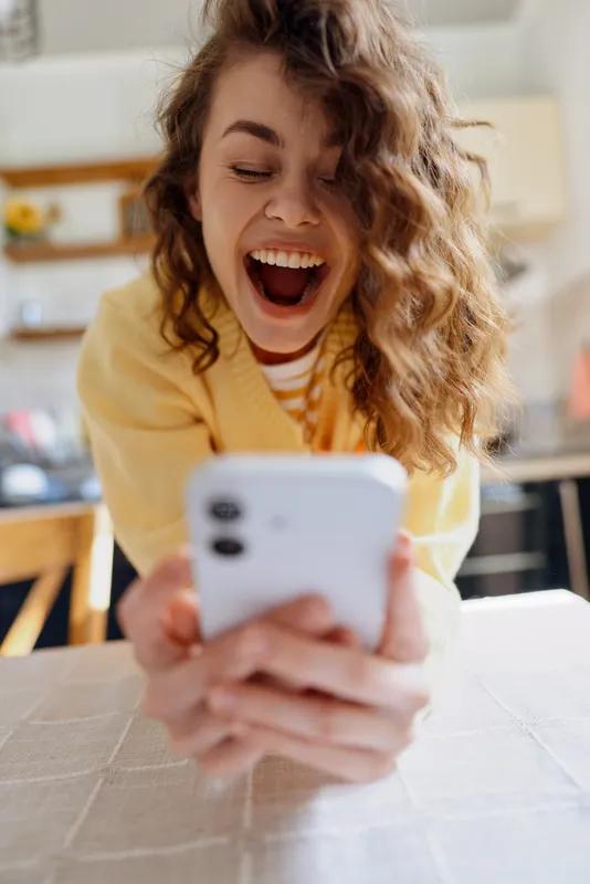 A young woman with curly hair laughs while looking at her phone in a cozy home setting, capturing a casual lifestyle moment with warm, genuine joy.