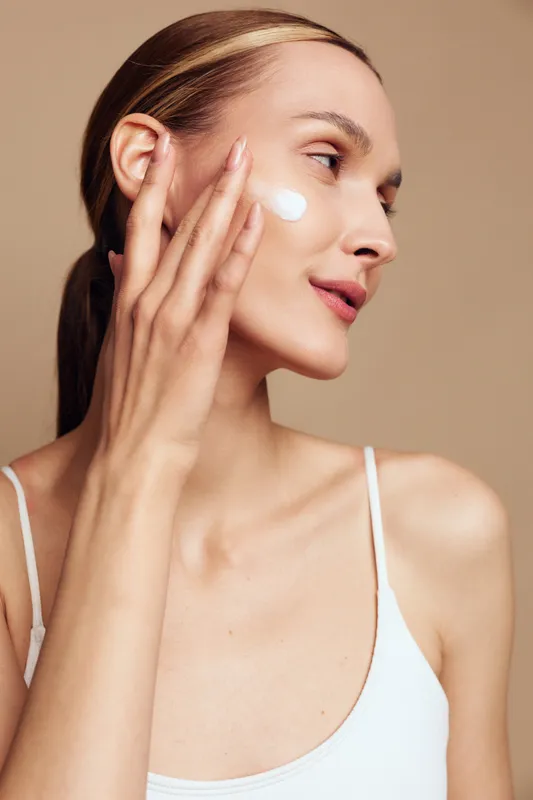 Young woman with clear skin applying white face cream on cheek. Close-up portrait on beige background. Concept of skincare, beauty routine, self-care, and natural wellness.