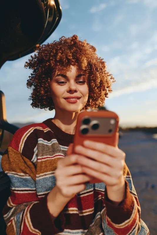 Woman lifestyle outdoors at sunset, curly hair woman in striped sweater holds smartphone for selfie, smiling softly, camera lens in foreground, warm evening atmosphere along open road
