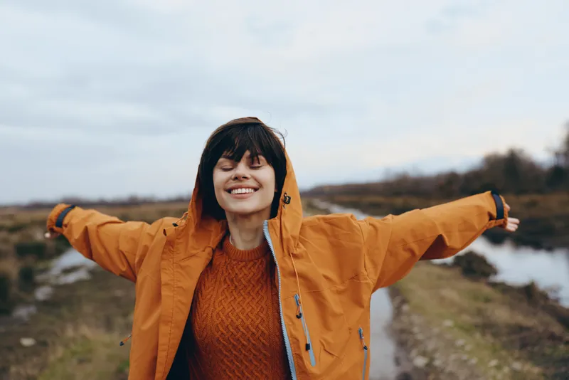 Smiling woman in orange jacket and hood outdoors on a riverbank, arms open in a cheerful lifestyle moment, autumn landscape with soft warm light near water, casual travel portrait