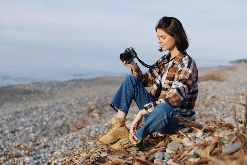 Woman with camera on a pebble beach, seated in casual attire, exploring photography by the sea. Windy, light, and relaxed vibes capture outdoor creativity.
