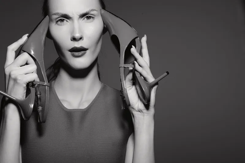 Portrait of a white woman with light skin holding high heel shoes near her face. Studio shot with dramatic lighting and neutral background. Concept of fashion and beauty.