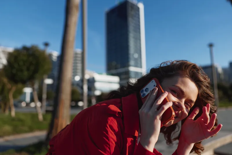 Young woman in a red jacket talks on a phone outdoors, sunny urban park scene, joyful expression, bright colors and casual pose