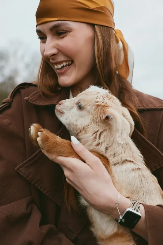 Young woman in a brown jacket smiles warmly while holding a baby goat close to her chest outdoors on a cloudy day, showing a tender moment of connection and care.