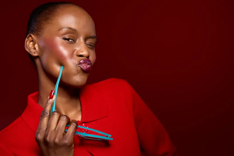 Portrait of a dark skinned African woman with glossy lips wearing red jacket holding blue sunglasses on red background. Concept of beauty, fashion, and confident female model expression.