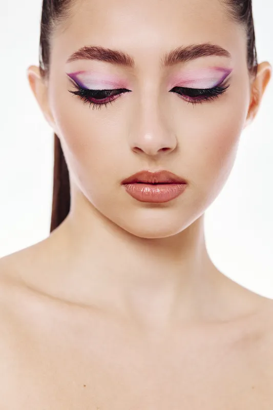 Closeup portrait of a young woman with creative purple and pink eyeshadow makeup, glossy nude lips, and perfect skin on a white background. Concept of beauty and fashion trends.