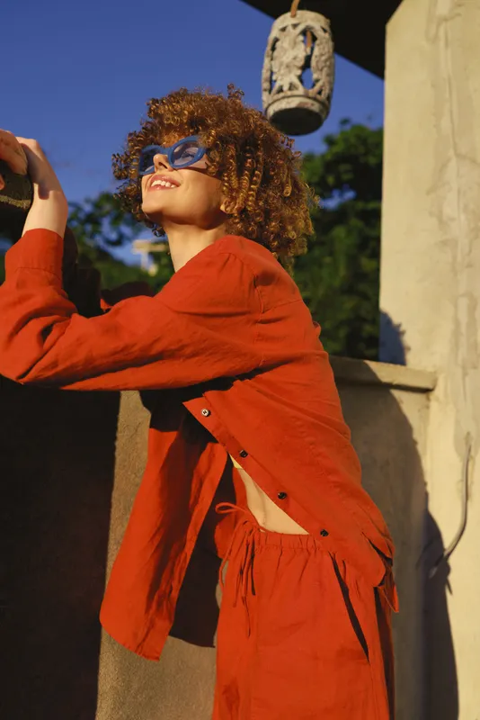 Cheerful woman outdoors, leaning on a sunlit railing, wearing orange cardigan, curly hair, sunglasses, relaxed pose, warm colors, carefree mood, stylish lifestyle moment in daylight