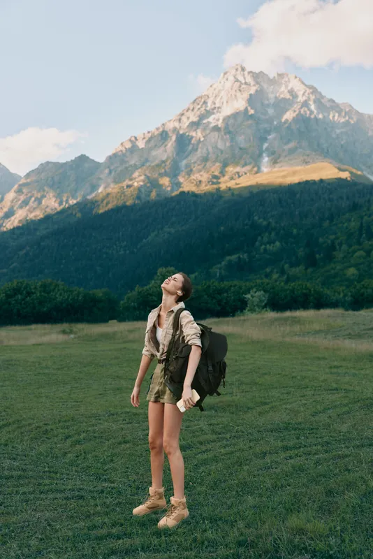 Young woman in hiking attire stands in a wide grassy meadow, gaze lifted toward rugged mountains behind, a sense of adventure