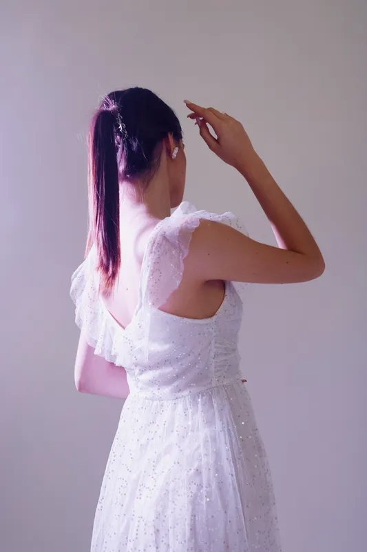 Young woman with long hair in a white lace dress poses with her back to the camera in soft light. Concept of beauty, fashion, and elegance.