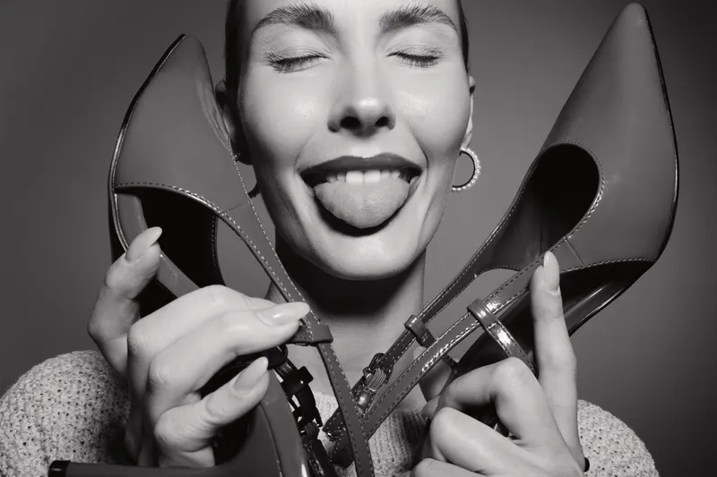 Happy white woman with light skin holding high heels near face shows tongue with closed eyes. Concept of fashion, fun, and playful female expression in studio black and white portrait.