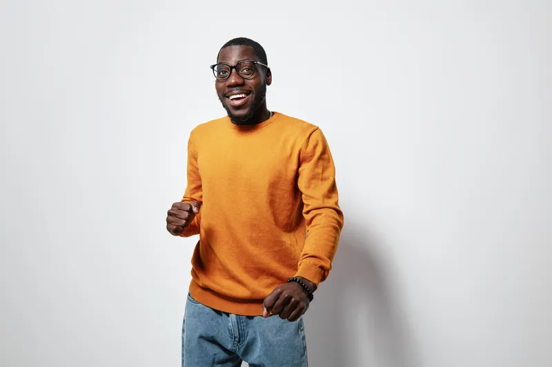 African American man with dark skin wearing glasses and orange sweater smiling and posing against white background. Casual style, happy expression and modern look.