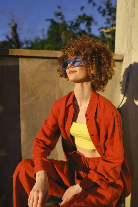 A stylish woman sits outdoors in bright sunlight, wearing a red suit and yellow top, posing confidently. A vivid lifestyle portrait highlighting fashion, color, and personal style