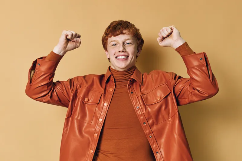 Happy young man with red hair wearing orange shirt and jacket smiling and raising fists on beige background with excitement and positive energy.