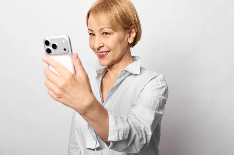 Senior woman smiles holding smartphone selfie gesture on neutral background isolated close up portrait, blonde hair, communication technology, casual indoor