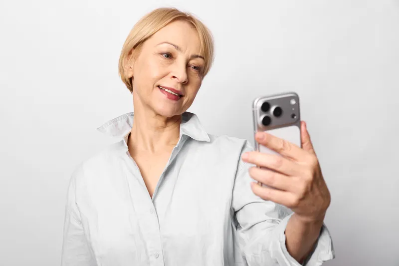 Senior woman takes a selfie with smartphone in a studio portrait, smiling, white background, using mobile technology for communication, indoors, relaxed calm closeup.