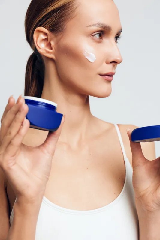 A young woman in a white tank top applies cream on her cheek, holding a blue jar in one hand and the lid in the other. The background is plain white, emphasizing skincare and beauty.