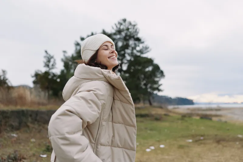 Woman in a warm beige jacket and knit beanie outdoors, enjoying the fresh breeze with a bright smile. Open landscape, bare trees, and distant water create a calm winter mood.